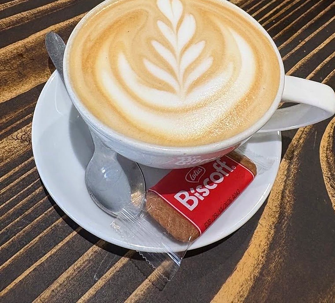A latte with leaf latte art on a wood-grain table, a Lotus Biscoff biscuit on the saucer