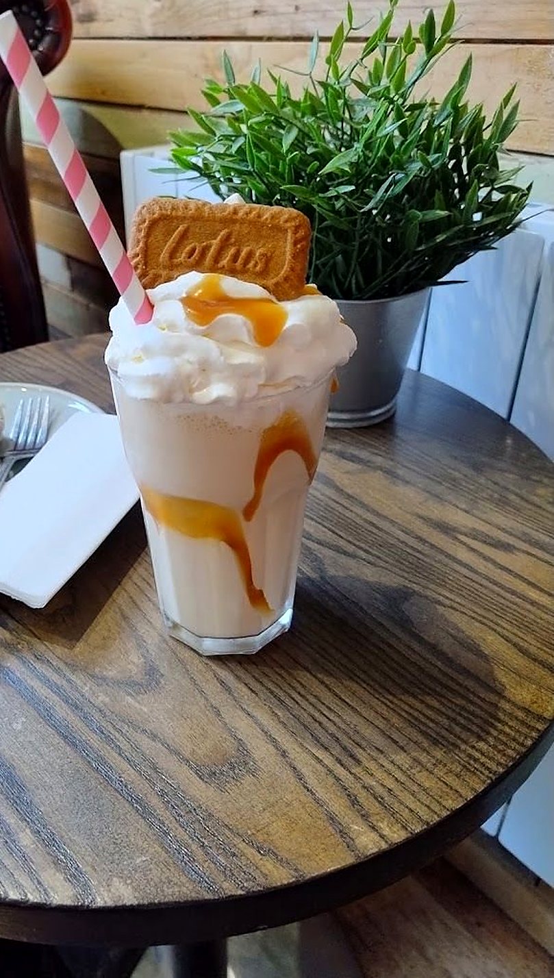 A milkshake topped with whipped cream, caramel sauce and a Lotus biscuit, with a striped paper straw and a slice of sponge cake